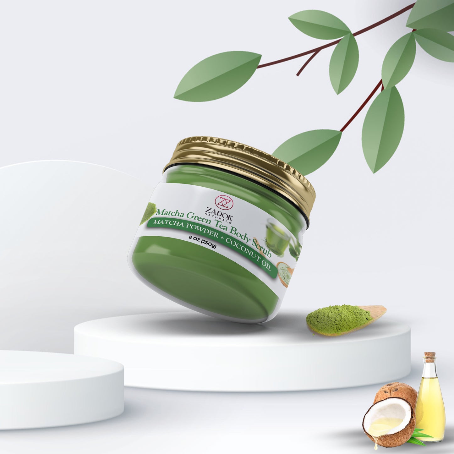 A jar of Matcha Green Tea Scrub with a green lid, accompanied by a coconut oil droplet and a piece of jojoba oil, set against a white background with green leaves in the background.